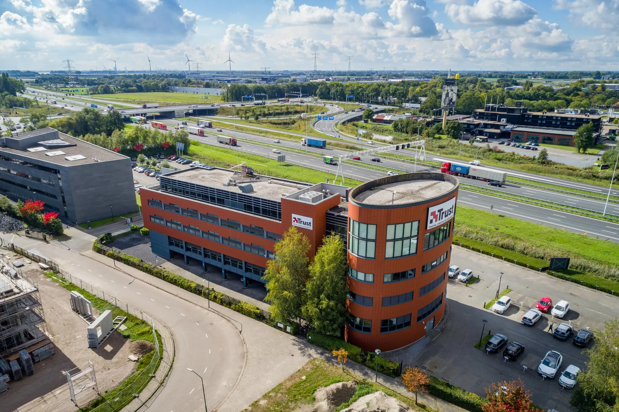 Luchtfoto van het kantoorgebouw van Trust aan de Laan van Barcelona, gelegen naast een snelweg met verkeer en omringd door zakelijke panden en landschap.